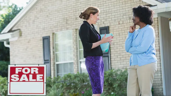 A real estate agent standing in front of a house with a FOR SALE sign in the yard, talking with an African American woman who could be the homeower, or a potential buyer.