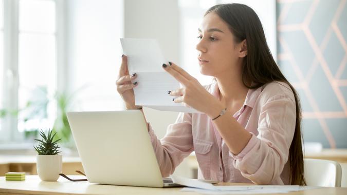 Concentrated female millennial worker reading contract, getting acquainted with agreement terms and conditions, woman employee considering document while working at laptop in modern office.