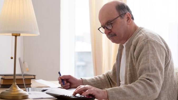 Senior man sitting with paperwork and using calculator while counting money stock photo