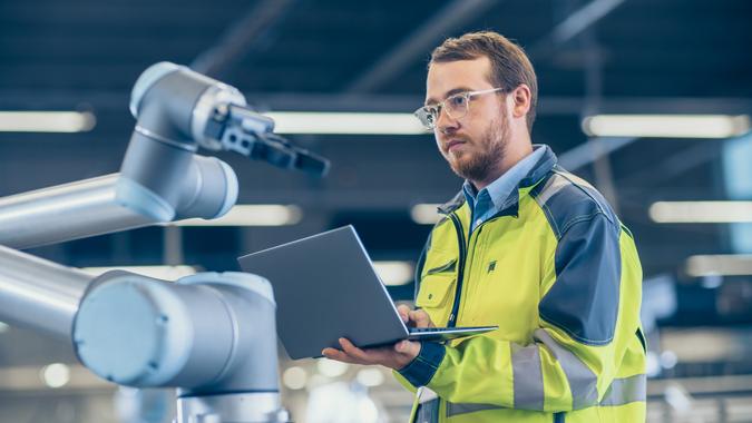 At the Factory: Automation Engineer Uses Laptop for Programming Robotic Arm.