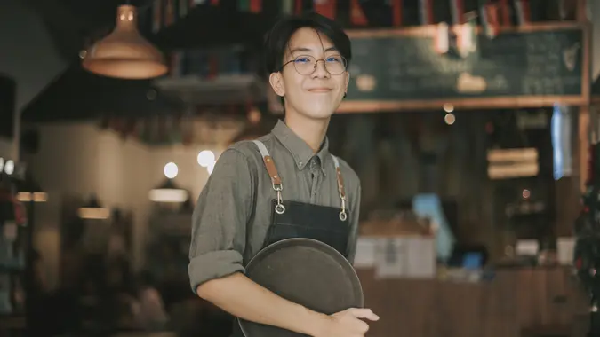 rear view asian chinese teenage boy waiter tying up apron getting read to work at cafe opening.