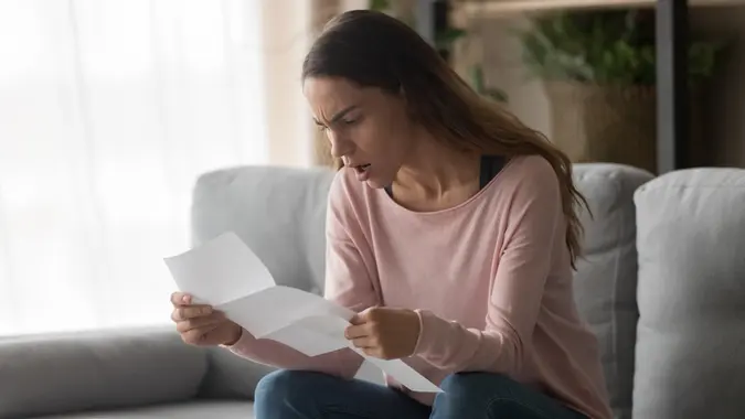 Angry woman sitting on couch at home holding letter reading awful news about company job refusal feels frustrated negative emotions face expressions, college admission or bank loan rejection concept.