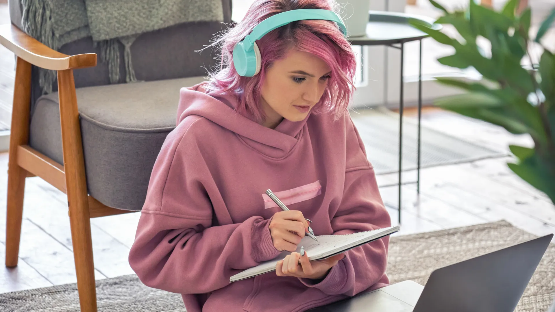 Focused hipster teen girl school college student pink hair wear headphones write notes watching webinar online video conference calling on laptop computer sit on floor working learning online at home.