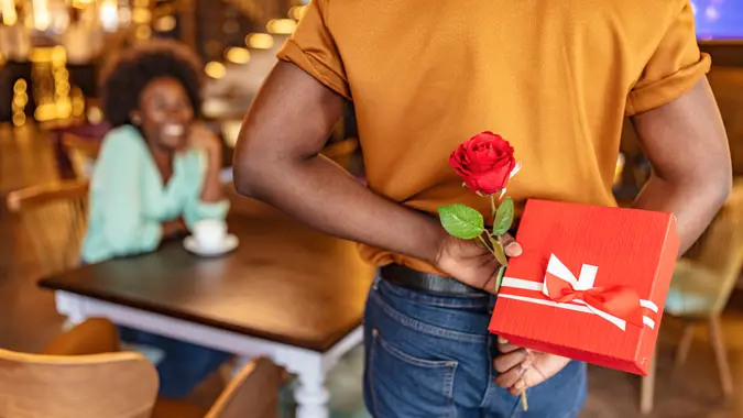 Man hiding red rose behind his back and gift for his smiling girlfriend while sitting in restaurant.