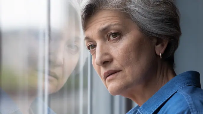 Close up head shot frustrated middle aged 60s woman standing near window, looking in distance, feeling depressed indoors.