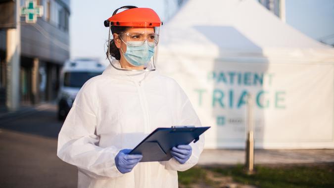 Paramedic wearing personal protective equipment PPE holding folder standing in front of ICU hospital isolation rt-PCR drive thru testing site, COVID-19 pandemic outbreak crisis, worried exhausted staff.