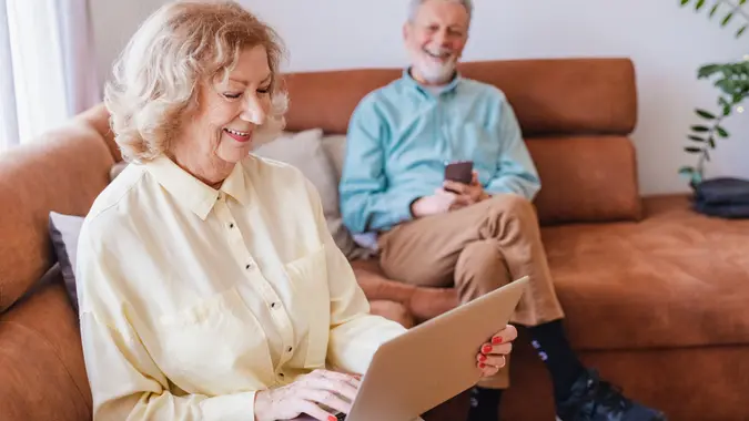Senior woman is using laptop at home, she is in the living room and her husband is in background.