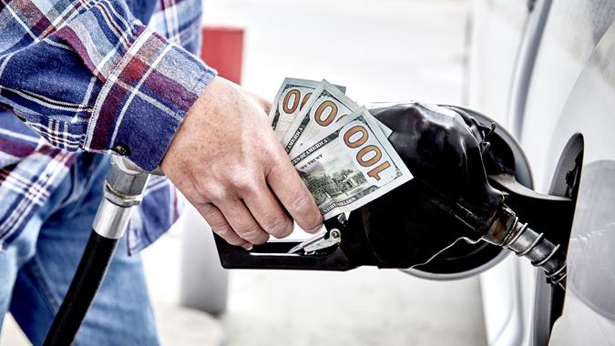 Man's hand holding three hundred US dollars and gas nozzle while  pumping gas into parked vehicle.
