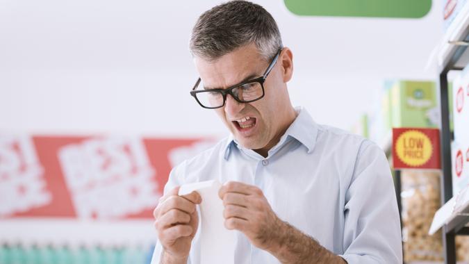 Shocked man doing grocery shopping at the supermarket and checking a long receipt, lifestyle and budgeting concept.
