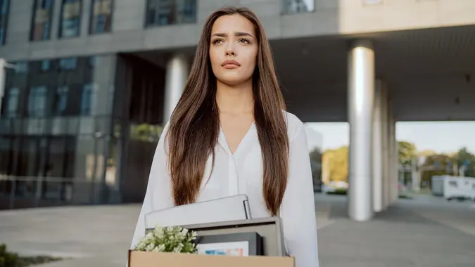 Woman stands in front of glass modern building of corporation office where she worked dismissal from position girl with sad uncertain face looks ahead holding box with packed things unemployment stock photo