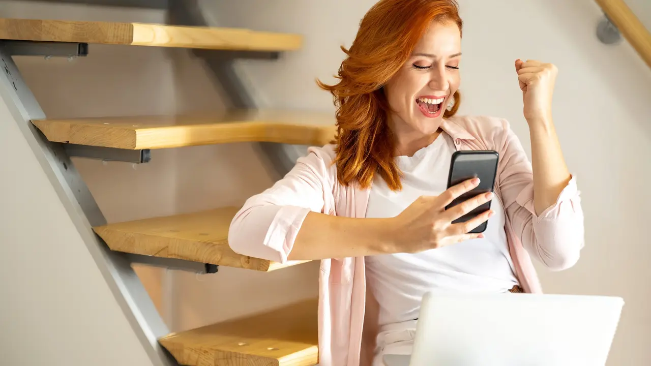 Portrait of euphoric young woman holding phone reading good news, while sitting over staircase at home. stock photo
