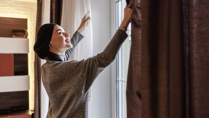 Young beautiful Muslim woman is opening blinds on a window in a living room. stock photo
