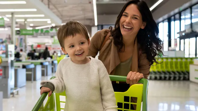 Beautiful mother pushing cart with her toddler inside with purchased groceries leaving the supermarket and facing camera smiling stock photo