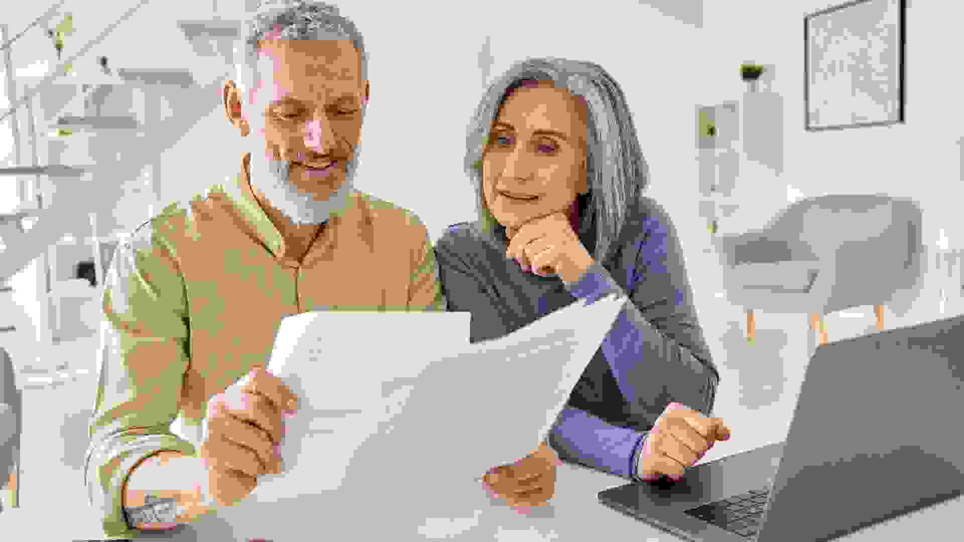 Middle aged senior old couple holding documents reading paper bills paying bank loan online, calculating pension fees, payments, taxes, planning family retirement money finances using laptop at home.