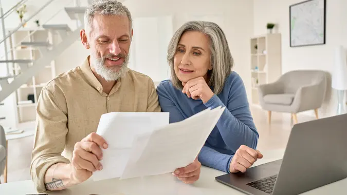 Middle aged senior old couple holding documents reading paper bills paying bank loan online, calculating pension fees, payments, taxes, planning family retirement money finances using laptop at home.