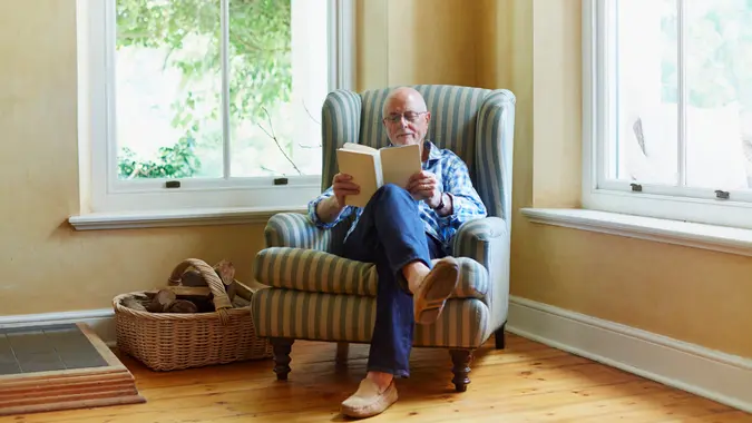 Full length of senior man reading book while relaxing on chair at home.