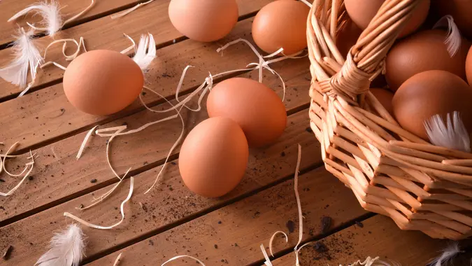 Freshly picked eggs in wicker basket on wooden table with straw and feathers.