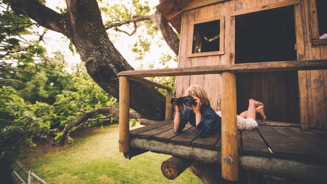 Young boy lying on the porch of a treehouse using binoculars to spy on something in the distance with leaves and a lush green grassy park in the background.