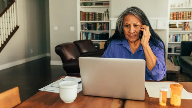 A senior aged woman sits at her kitchen table while paying medical bills, talking with her doctor, and updating medicine prescriptions.