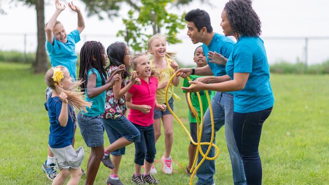 A group of children are jumping in excitement as they are getting ready with their camp counsellors to play a game of tug-of-war at summer camp.