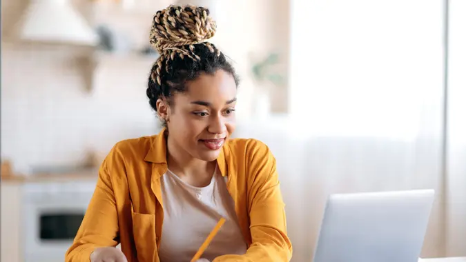 Focused cute stylish african american female student with afro dreadlocks, studying remotely from home, using a laptop, taking notes on notepad during online lesson, e-learning concept, smiling.