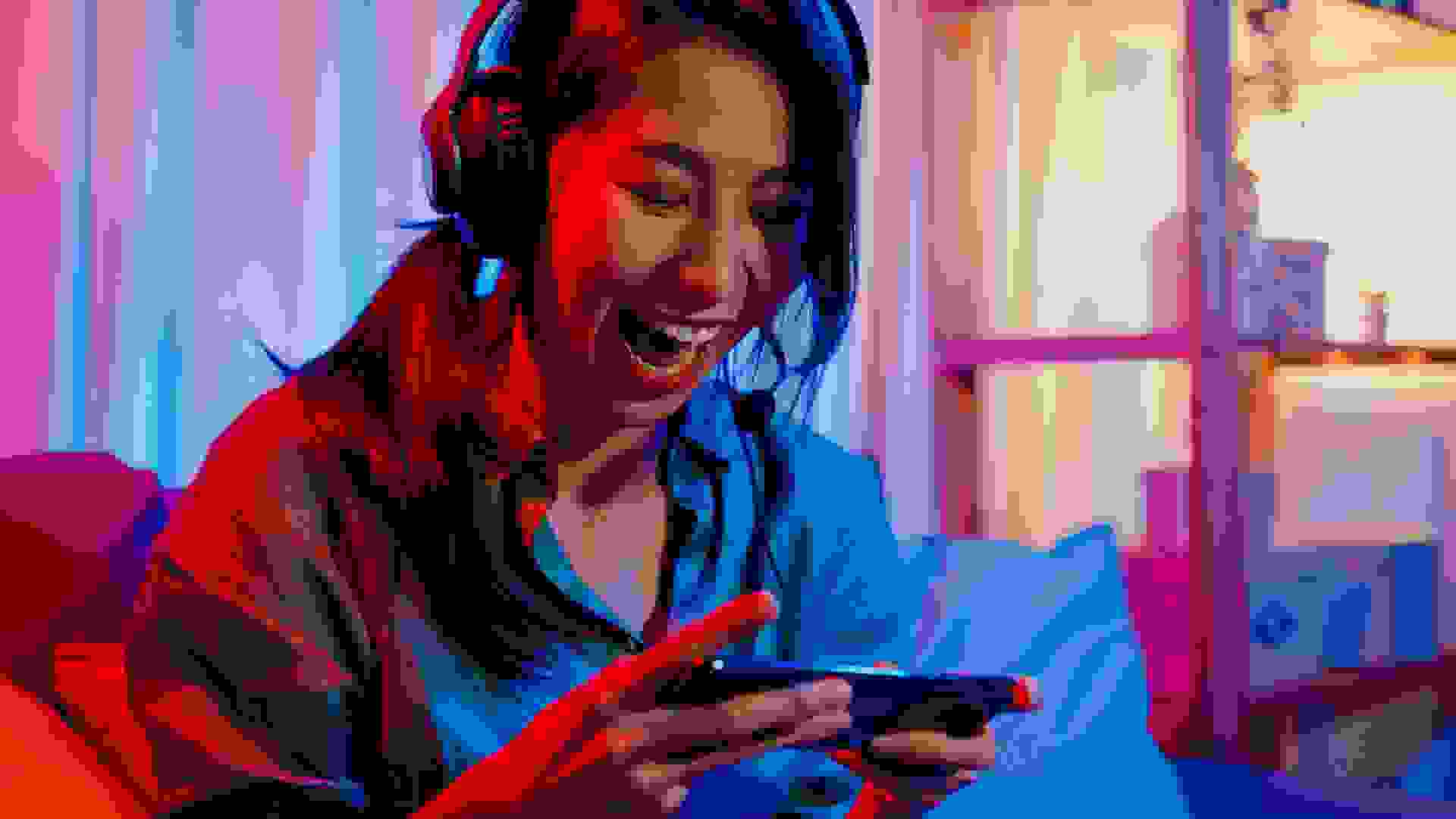 Happy asia girl gamer wear headphone competition video game online with smartphone excited talk with friend sit on couch in colorful neon lights living room at home.