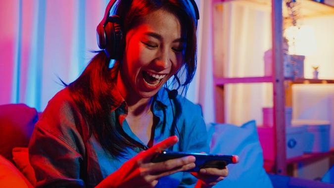 Happy asia girl gamer wear headphone competition video game online with smartphone excited talk with friend sit on couch in colorful neon lights living room at home.