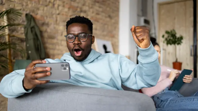 Young African American man relaxing at home in a loft apartment with his Caucasian female roommate, playing video games on a smart phone while sitting on a sofa and having fun.
