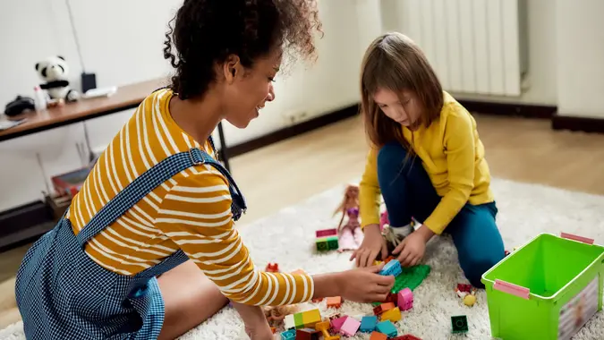 Have fun. Caucasian cute little girl spending time with african american baby sitter. They are playing with construction toys set, sitting on the floor stock photo
