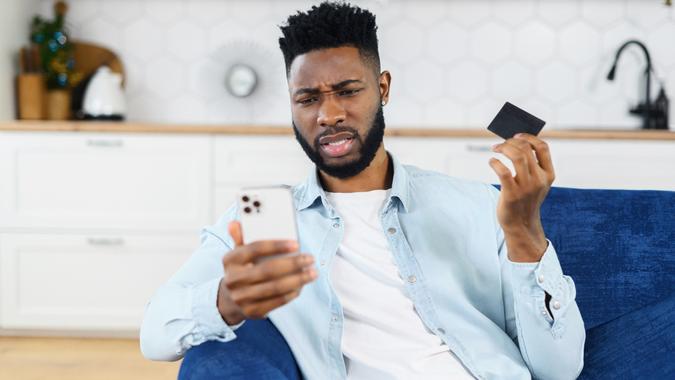 非洲裔美国人担心钱。 困惑的黑人拿着信用卡和智能手机，失望的脸耸了耸肩 库存照片