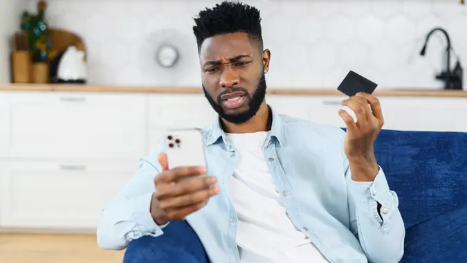 African-American worrying about money. Confused black man holding credit card and smartphone with disappointed face, shrugging stock photo