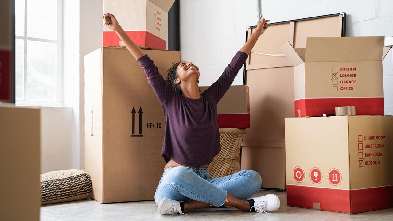 Happy young woman sitting in new apartment and raising arms in joy after moving in.