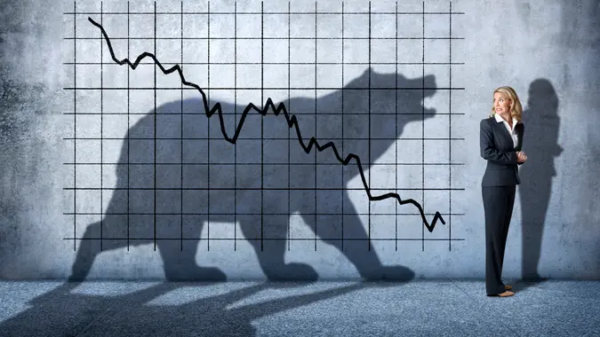 A businesswoman looks over her shoulder with concern at  a descending stock chart and an ominous shadow of a bear that is cast on the wall above her.