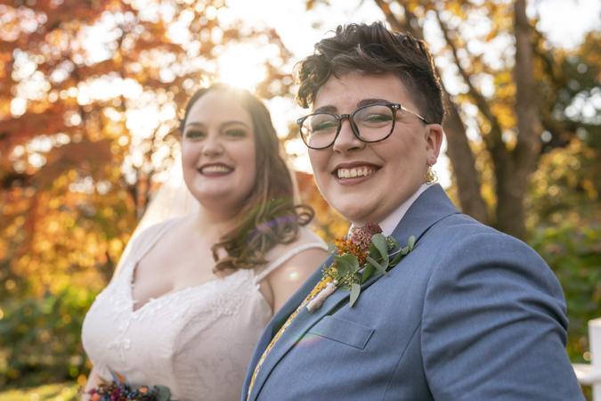 A young and beautiful lesbian couple smile directly at the camera while celebrating their marriage at an outdoor reception party on their wedding day.