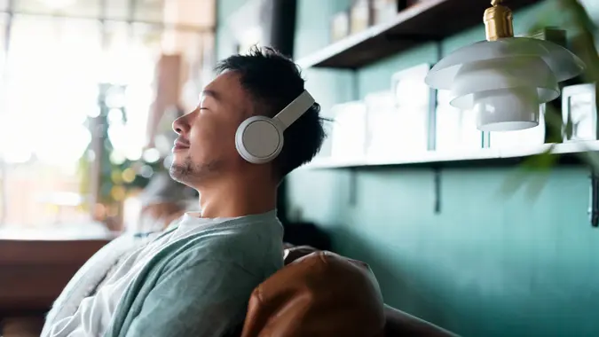 Young Asian man with eyes closed, enjoying music over headphones while relaxing on the sofa at home.
