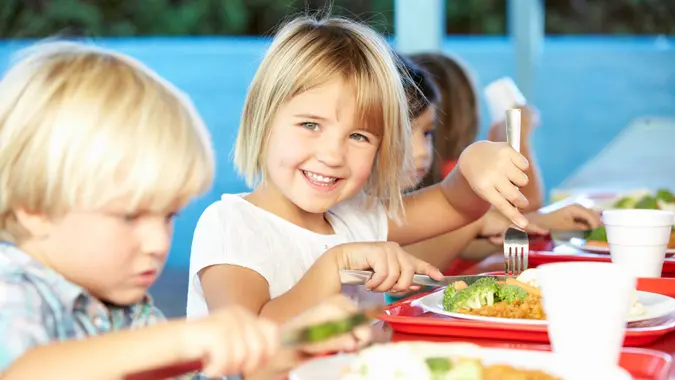 Elementary Pupils Enjoying Healthy Lunch In Cafeteria Sitting Down Smiling At Camera.