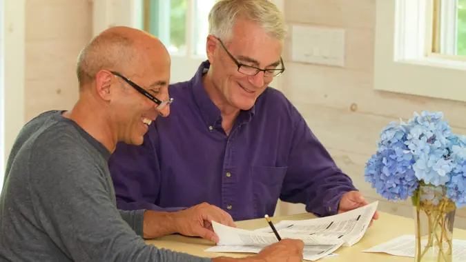 Senior gay male couple, smiling and affectionate, working at their kitchen table on financial documents such as wills, financial planning, or a contract for a house.