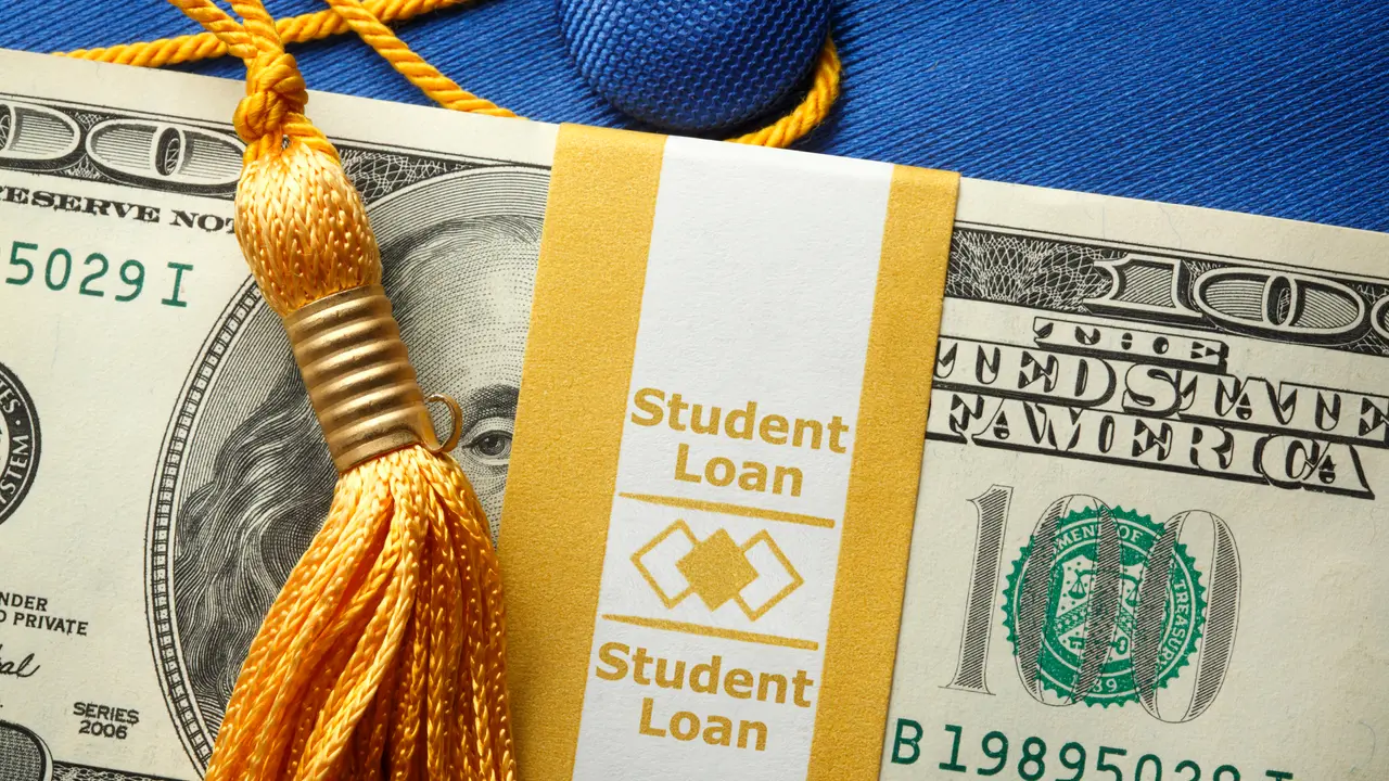 A stack of one hundred dollar bills in a money wrapper labeled "Student Loan" on top of a blue graduation cap.