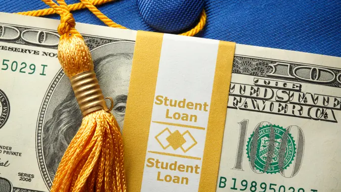 A stack of one hundred dollar bills in a money wrapper labeled "Student Loan" on top of a blue graduation cap.