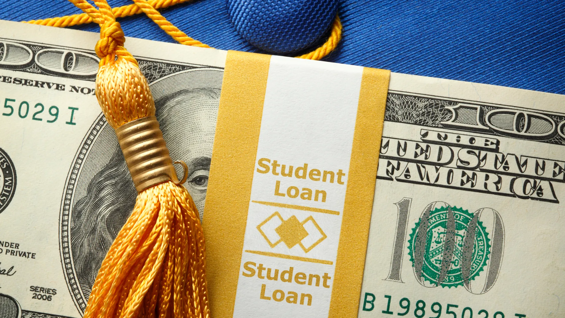一叠一百美元的钞票放在一个印有“学生贷款”字样的钞票包装里，上面放着一个蓝色的毕业帽。