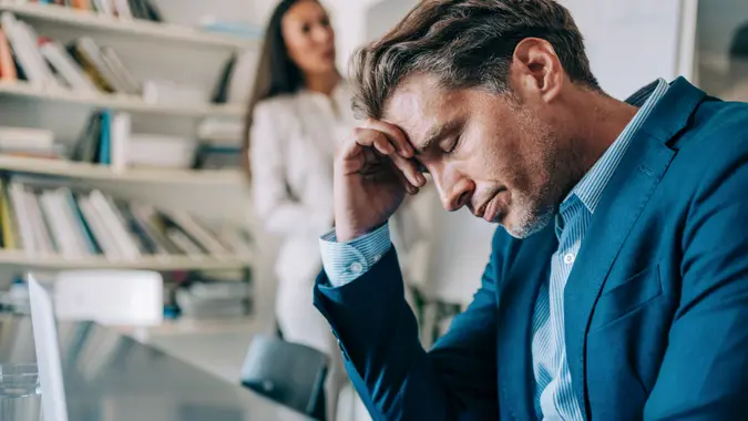 Overworked businessman sitting in the board room and holding head with hand while his female colleague making a presentation on whiteboard.
