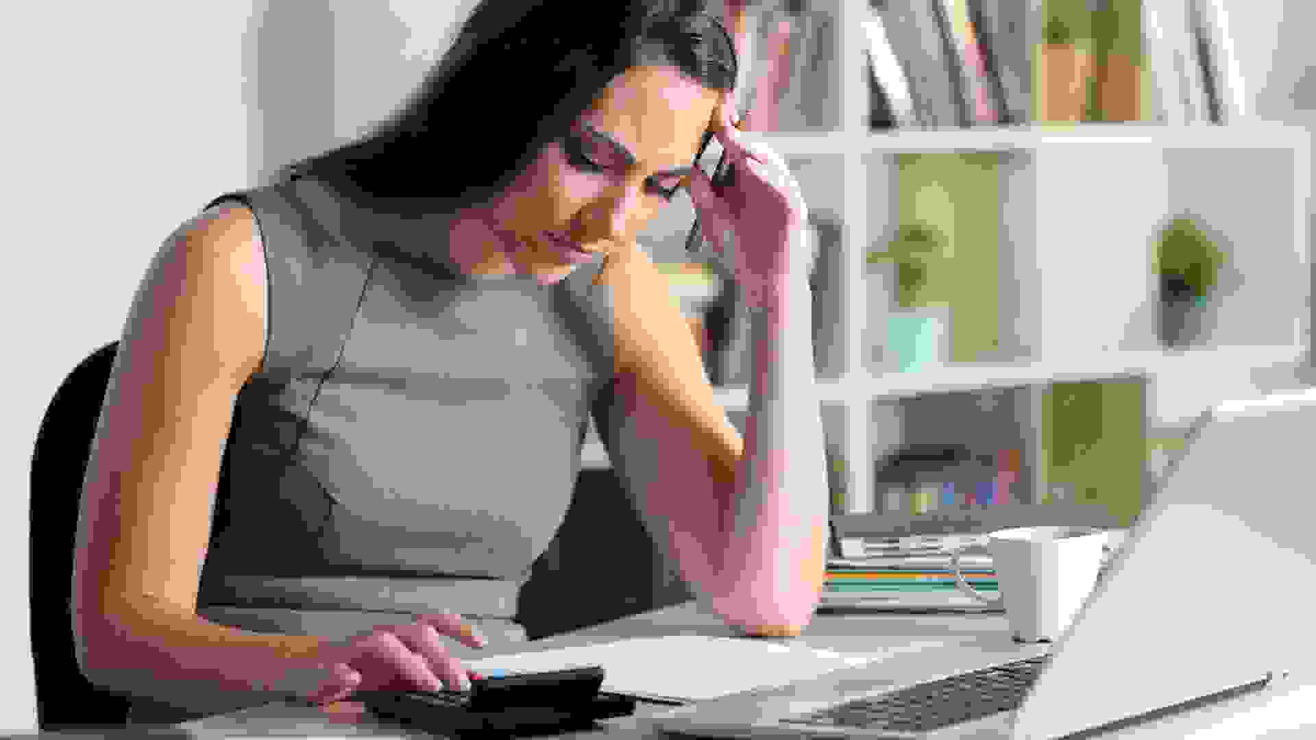 Worried bookkeeper woman calculating looking at calculator sitting on a desk at homeoffice.