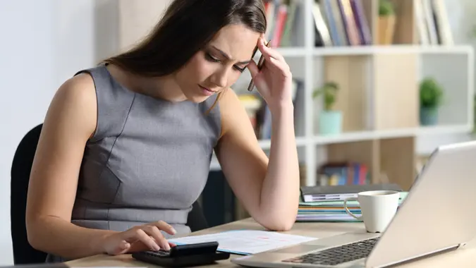 Worried bookkeeper woman calculating looking at calculator sitting on a desk at homeoffice.