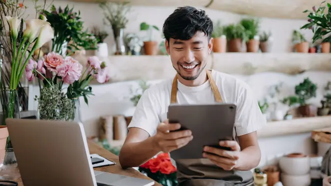 Asian male florist, owner of small business flower shop, using digital tablet while working on laptop against flowers and plants.