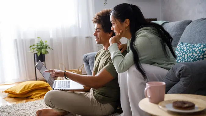 Wide shot of millennial couple lounging in their living room, planning out their finances and looking at their account via online banking.