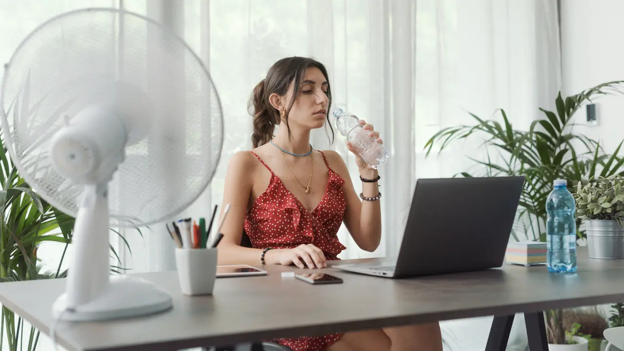 Woman sitting at desk at home and working with a computer, she is cooling herself with an electric fan and drinking water.