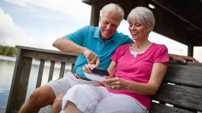 Happy senior couple depositing bank check through mobile phone on wooden bench near pond.