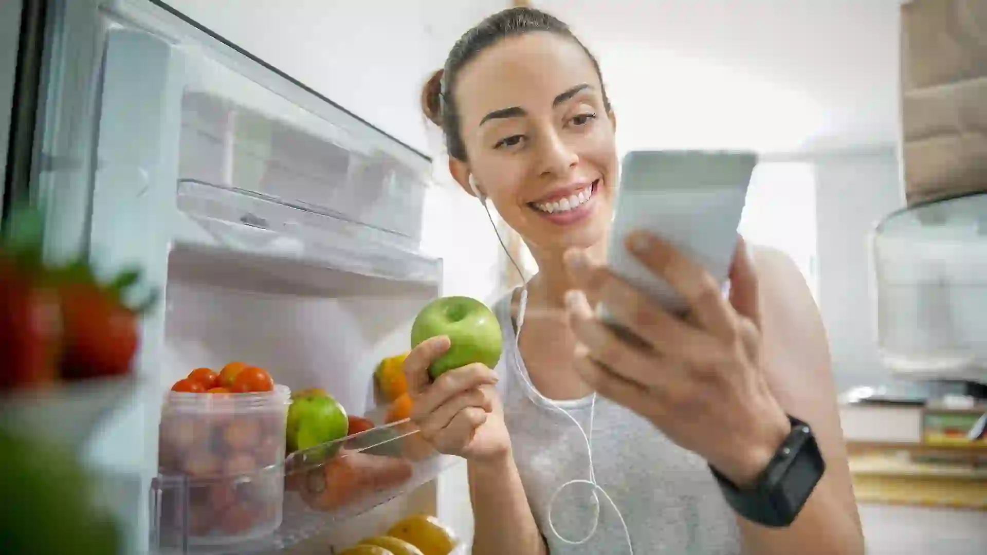 Happy woman at home grabbing food from the fridge while watching videos online stock photo