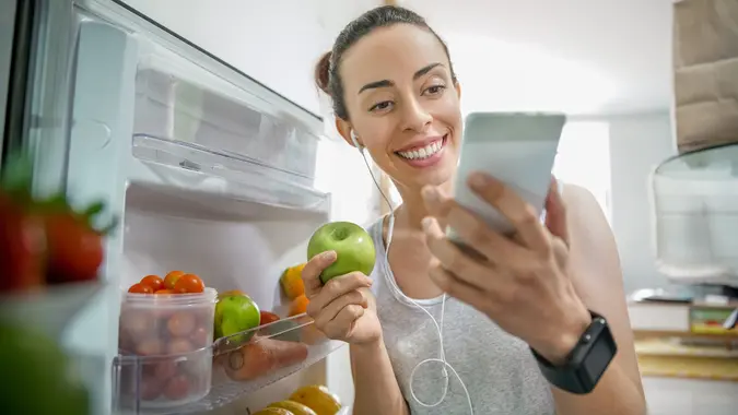 Happy woman at home grabbing food from the fridge while watching videos online stock photo