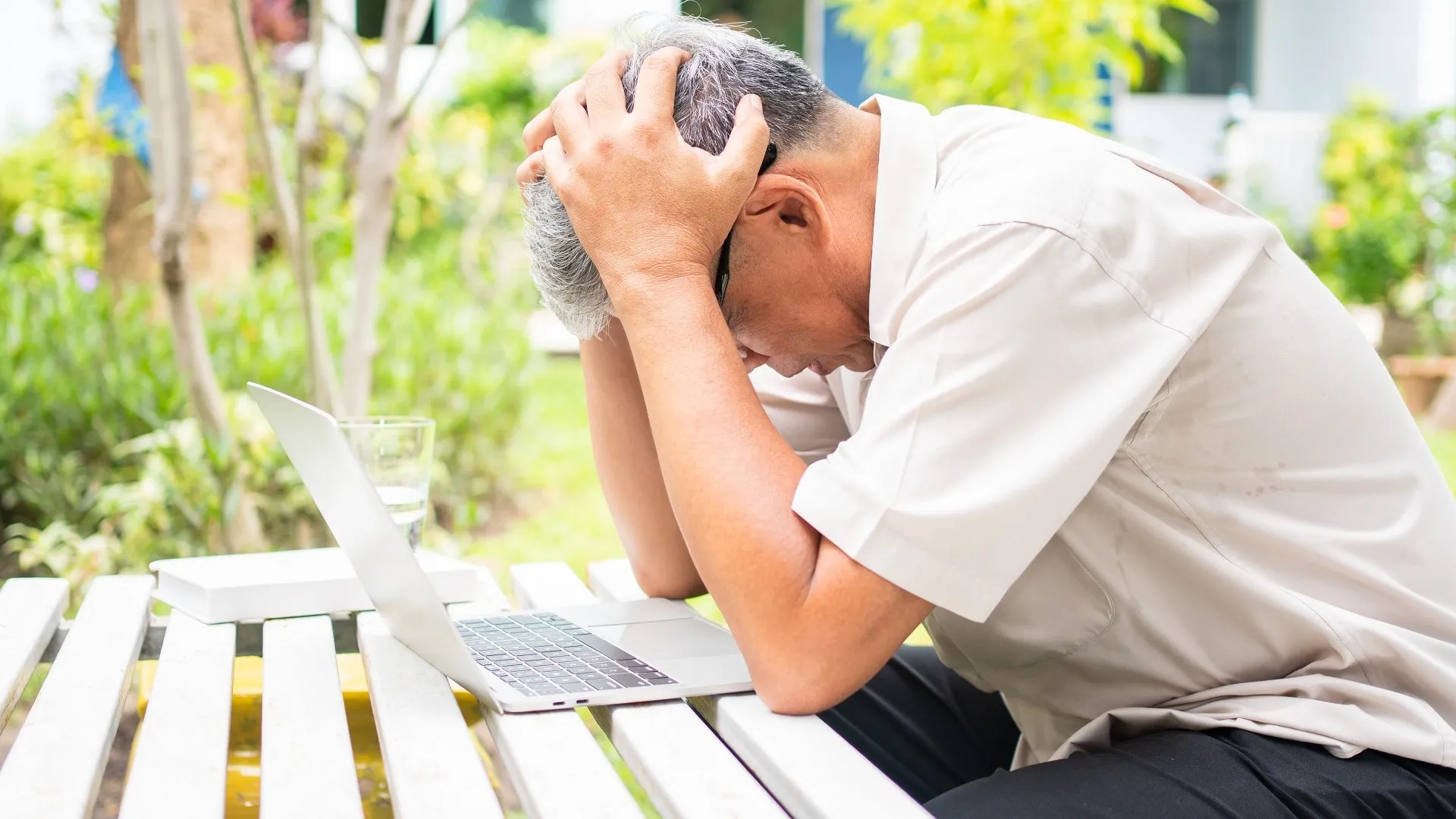 Portrait of an old elderly Asian man holds the head with his hand cause of stress after try to use a computer laptop in the backyard after retired. Concept of Ageism and Hobbies after retirement. stock photo